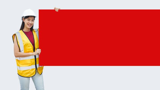 Eine asiatische Ingenieurin mit gelbem Westentuch und weißem Helm stand mit einem roten Papierplakat mit leerem Platz für Text auf isoliertem Hintergrund. - Foto, Bild