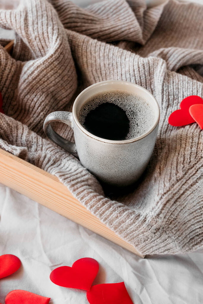Керамічна сіра чашка з чорною кавою в затишній атмосфері на бежевій зіпсованій тканині ліжка і багато червоних сердець декору. сюрприз для коханої, ідея дарування для дня валентинки, вертикальний зміст - Фото, зображення