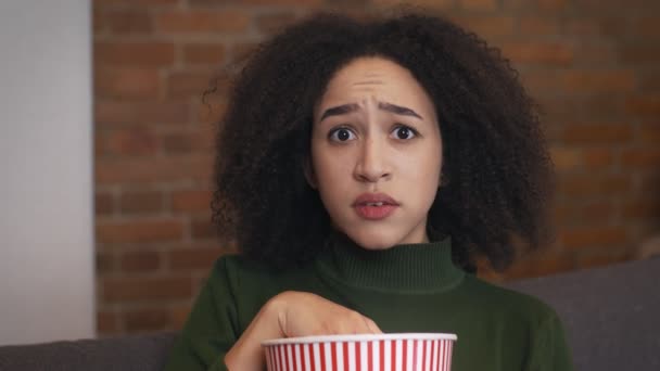 Young emotional black woman watching tv at home and eating popcorn, feeling frightened and afraid, close up portrait - Footage, Video