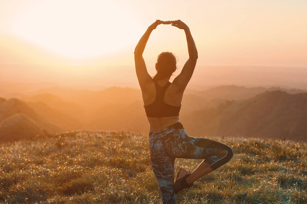 Una vista posteriore di una donna in piedi su una gamba mentre pratica la posa albero yoga mentre guarda il tramonto - Foto, immagini