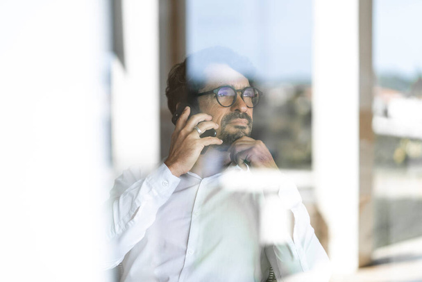 Un uomo con gli occhiali che fa una telefonata da cellulare seduto a guardare dalla finestra. Ritratto maschile di successo che lavora da casa - Foto, immagini