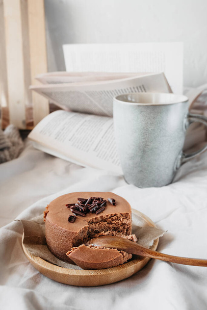 Небольшой круглый шоколадный чизкейк без выпечки на многоразовой бамбуковой тарелке на уютном бежевом одеяле с чашкой кофе и книгой. поддержание психического здоровья, время, проведенное в удовольствии. вертикальная, избирательная направленность - Фото, изображение