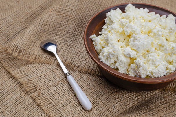 Close-up of fresh cottage cheese in a clay bowl. A full bowl of curd and a spoon on a sack. Ready to eat. Healthy food. Selective focus. - Φωτογραφία, εικόνα