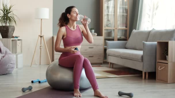 Pełne ujęcie radosnej białej kobiety ubranej w strój do jogi, siedzącej na fitballu w środku salonu, pijącej wodę z butelek sportowych i uśmiechniętej - Materiał filmowy, wideo