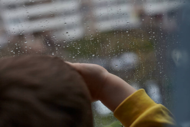 Ένα μικρό αγόρι φαίνεται θλιβερό έξω από το παράθυρο. Η έννοια του βροχερού καιρού. Το παιδί μένει σπίτι λόγω της βροχής. - Φωτογραφία, εικόνα
