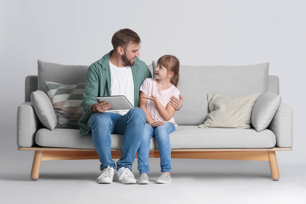 Father and daughter with tablet computer on sofa against light background - Фото, зображення