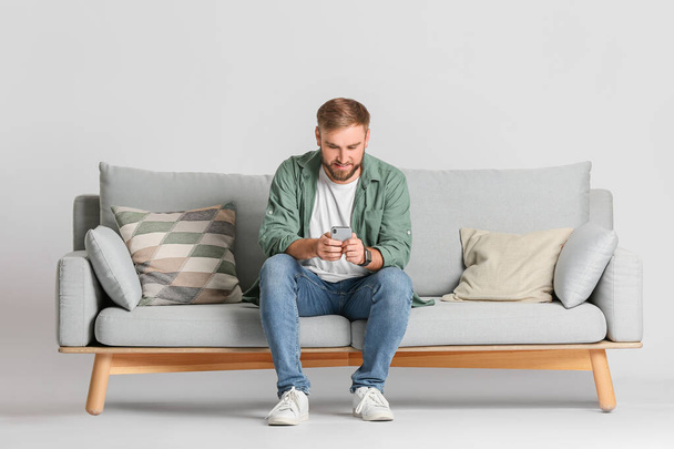 Young man with mobile phone on sofa against light background - Photo, image
