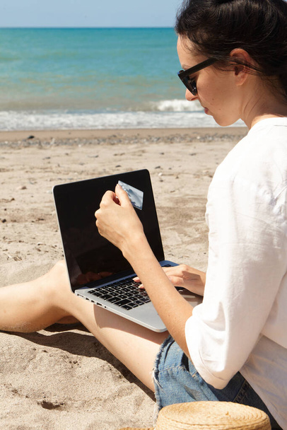 Nuori nainen käyttää kannettavaa tietokonetta rannalla. Loma elämäntapa viestintä. Hänellä on pankkikortti käsissään. Verkkokaupan tai verkkokaupan käsite - Valokuva, kuva