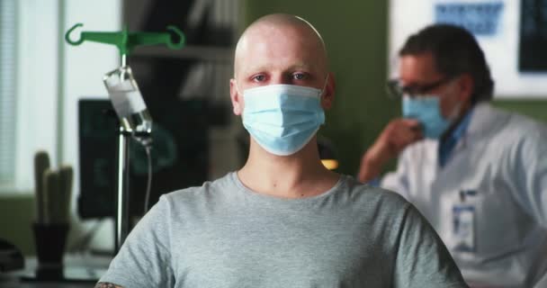 Лысый мужчина в маске смотрит в камеру во время химиотерапии - Кадры, видео