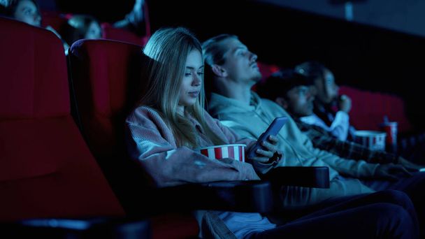 Beautiful young woman using her phone for texting while missing boring movie at the cinema - Фото, изображение