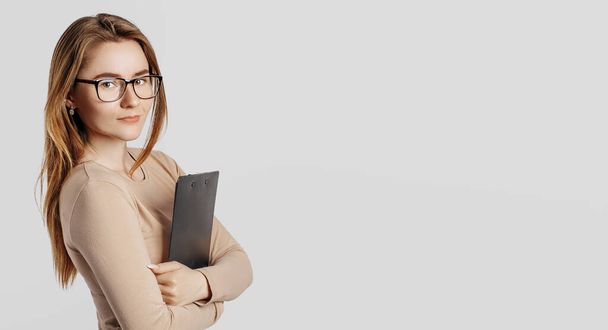 Beautiful young business woman wearing glasses isolated on gray background. Achieving career wealth business concept. Mockup copy space. Hold a clipboard with documents - Φωτογραφία, εικόνα