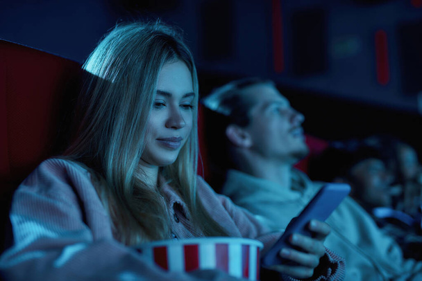 Porträt einer schönen jungen Frau, die lächelt und ihr Handy benutzt, während sie einen langweiligen Film im Kino verpasst - Foto, Bild
