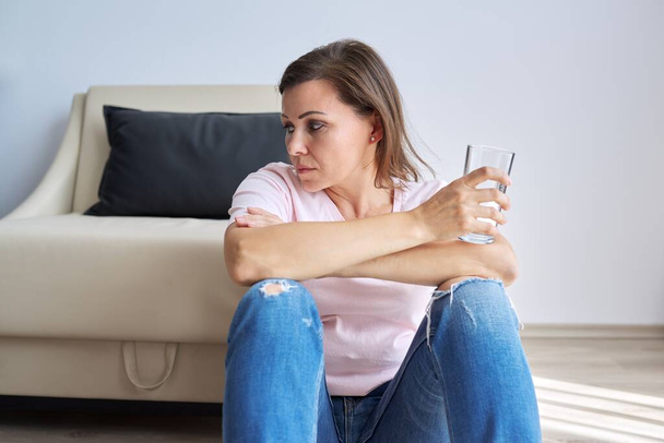Sad middle aged woman sitting at home on the floor with glass of water - Foto, immagini