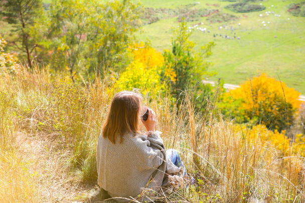 Beautiful girl drinking coffee in autumn nature. A cup with hot drink in the mountains. Cosy mood. Red and yellow leaves. Stylish blanket on woman's shoulders. View from the hill. - Photo, image