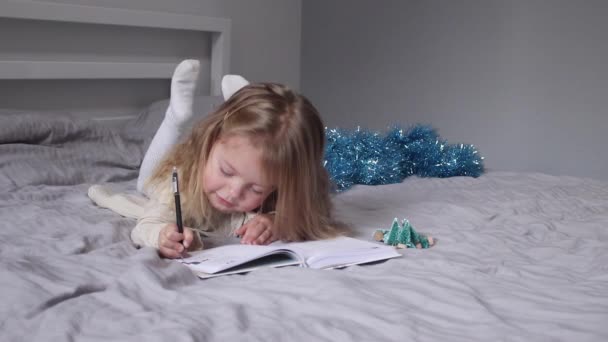 bájos kislány levelet ír a Télapónak az ágyon a szürke hálószobában. Újév, gyermekkor, karácsony. FullHD felvétel - Felvétel, videó