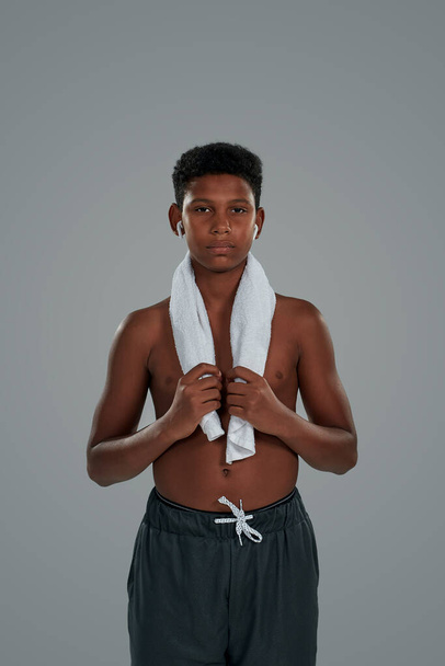 Resting after workout. Half naked teenage african boy with towel on shoulders wearing wireless earphones looking at camera while standing against grey background - 写真・画像