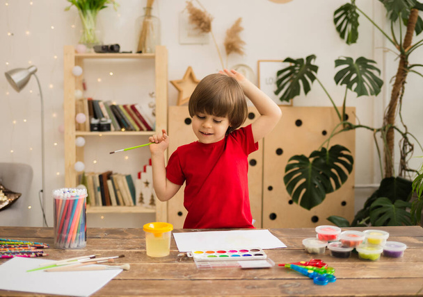 Mutlu küçük çocuk bir fırçayla çiziyor ve ahşap bir masanın üzerine kırtasiyeyle beyaz bir kağıt çiziyor. Çocuğun yaratıcı gelişimi - Fotoğraf, Görsel