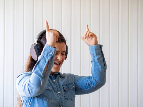 Γυναίκα ακούει μουσική και χορεύει με ασύρματα ακουστικά στο σπίτι. Διασκεδαστική δραστηριότητα κατά την καραντίνα. Καυκάσια γυναίκα σε denim χαρούμενη και διασκεδάζουν σε ένα διαμέρισμα.  - Φωτογραφία, εικόνα