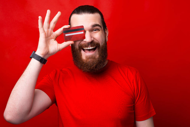 Веселий бородатий молодий чоловік тримає кредитну картку біля очей посміхаючись
 - Фото, зображення