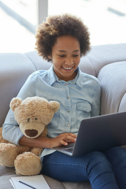 Löydä uusia etuja. Happy mixed race teini koulutyttö hymyilee opiskellessaan kannettavan tietokoneen avulla, istuu sohvalla kotona - Valokuva, kuva
