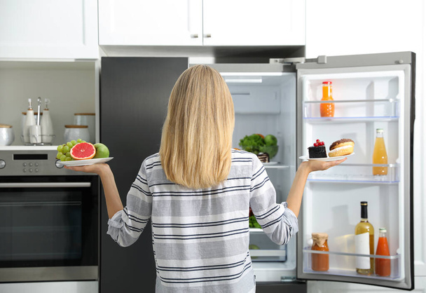 Choice concept. Woman holding plates with fruits and sweets near refrigerator in kitchen, back view - Foto, Imagem