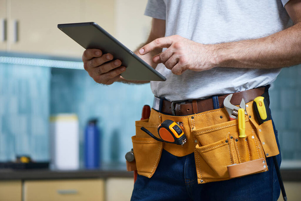 Cropped shot of young repairman wearing a tool belt with various tools using digital tablet while standing indoors - Фото, изображение