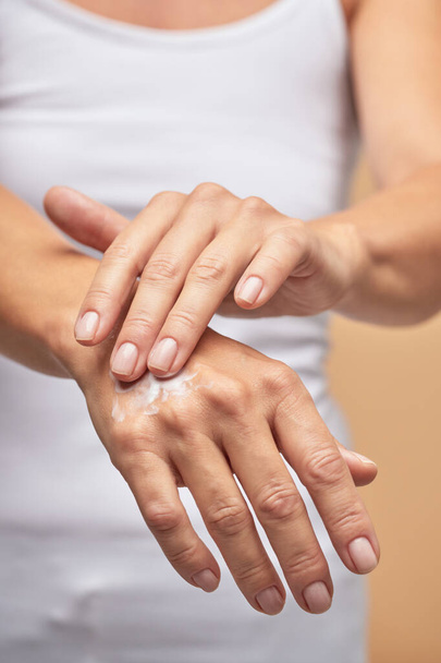 For dry skin. Vertical shot of a woman rubbing moisturizer on hands on beige background - Fotografie, Obrázek