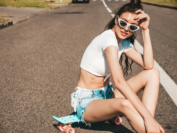 Young beautiful sexy smiling hipster woman in sunglasses.In white summer swimwear bathing suit.Positive female going crazy with blue penny skateboard.Model sitting on asphalt in the street - Foto, imagen