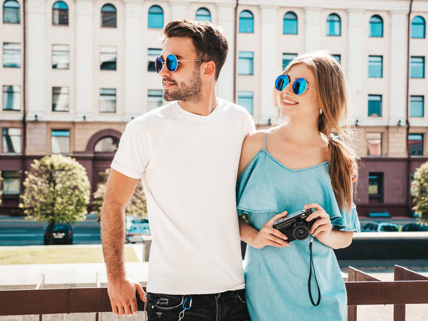 Smiling beautiful female and her handsome boyfriend.Woman in casual summer jeans dress.Happy cheerful family.Female having fun.Couple posing on the street background in sunglasses with retro camera - Photo, Image