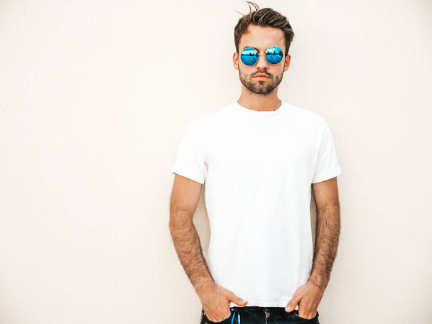 Portrait of handsome confident stylish hipster lambersexual model.Man dressed in white T-shirt. Fashion male posing on the street background near wall in sunglasses outdoors - Photo, image