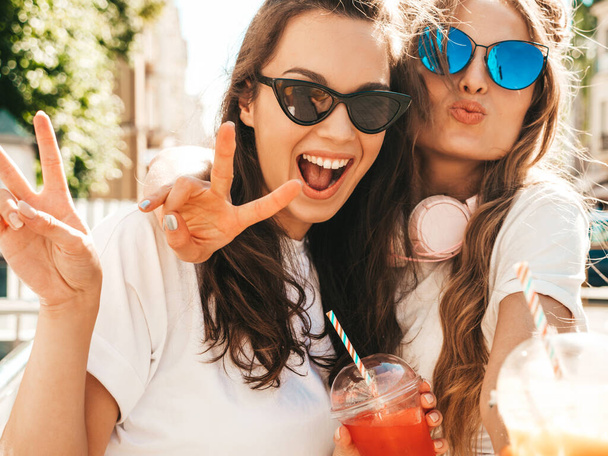 Two young beautiful smiling hipster female in trendy summer clothes. Carefree women posing outdoors.Positive models holding and drinking fresh cocktail smoothie drink in plastic cup with straw - Foto, afbeelding