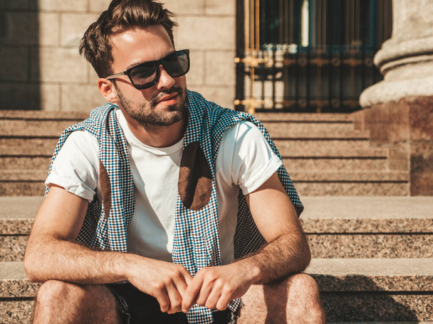 Portrait of handsome confident stylish hipster lambersexual model.Man dressed in white T-shirt. Fashion male sitting at the stairs on the street background in sunglasses - Foto, Bild