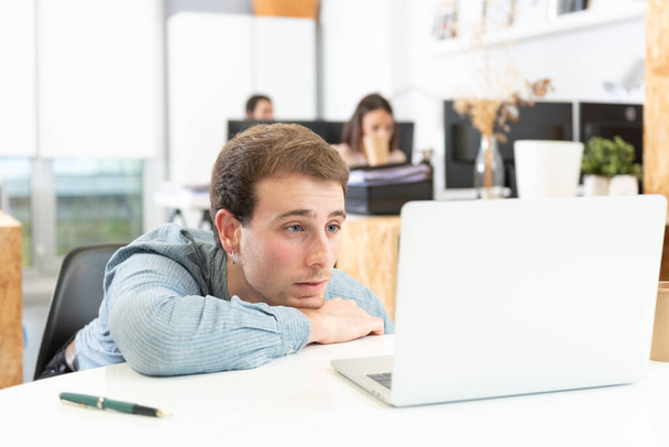 A bored and dispaired caucasian young man in front of his laptop in the office. - Photo, Image
