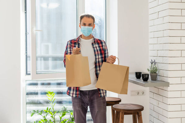 Young delivery man in medical mask on face holding paper bags in hands. Hipster man courier with shopping bags. Safe online delivery service. Coronavirus COVID-19 protection - Photo, image