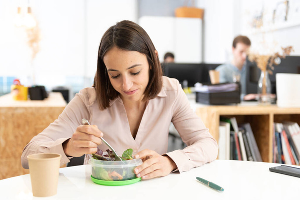 Zdrowe jedzenie w koncepcji miejsca pracy. Ładna latynoska kobieta je sałatkę w biurze.. - Zdjęcie, obraz