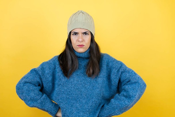 Νεαρή όμορφη γυναίκα φορώντας μπλε casual πουλόβερ και μάλλινο καπέλο σκεπτικός και νευρικός, αποδοκιμάζοντας έκφραση στο πρόσωπο με τα χέρια στη μέση - Φωτογραφία, εικόνα