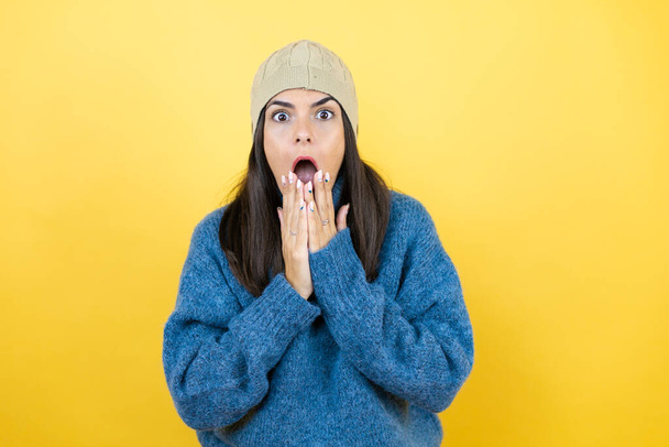 Young beautiful woman wearing blue casual sweater and wool hat shocked covering mouth with hands for mistake. secret concept. - Photo, Image