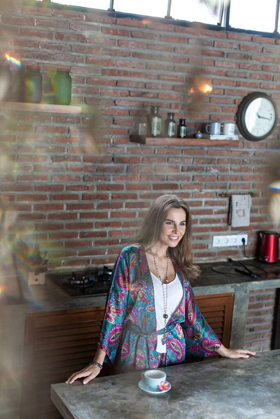 young beautiful slim woman with long brown hair in silk cardigan with purple and navy blue Indian patterns stay at table with blue tea cup in kitchen with break wall. through the glare - Foto, Imagen