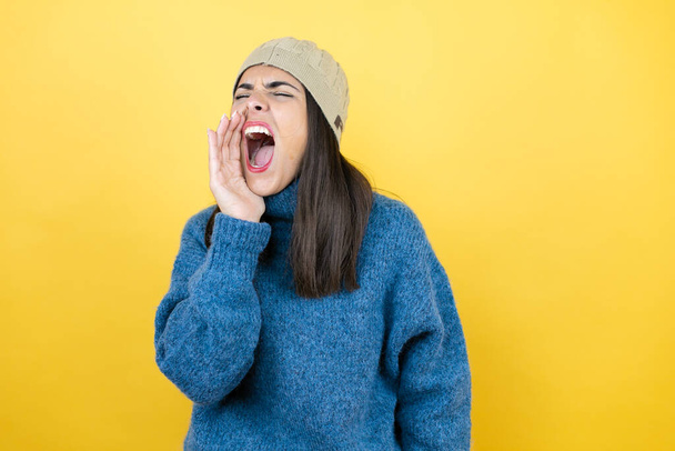 Young beautiful woman wearing blue casual sweater and wool hat shouting and screaming loud to side with hand on mouth - 写真・画像