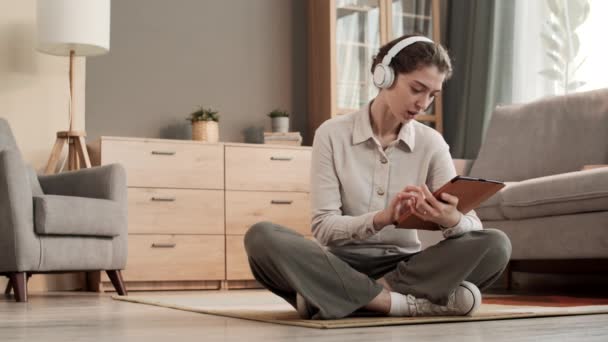 Πλήρης εικόνα της νεαρής Καυκάσιας γυναίκας που φοράει ασύρματα ακουστικά πάνω από το αυτί, κάθεται με τα πόδια σταυρωμένα στο πάτωμα στο σαλόνι, χρησιμοποιώντας τον υπολογιστή tablet, χαμογελώντας, δείχνοντας ευχαριστημένος - Πλάνα, βίντεο