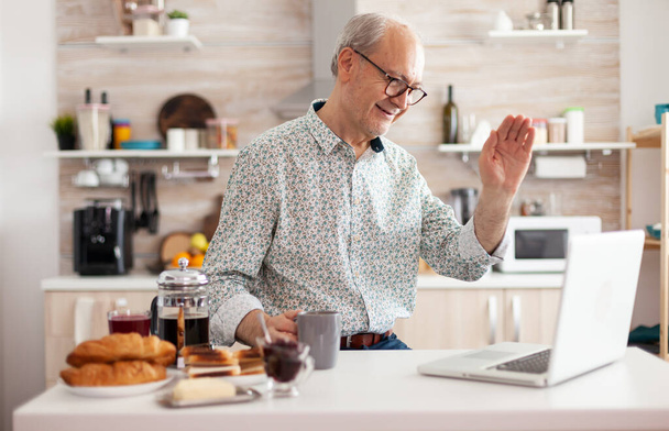 La vida cotidiana del hombre mayor en la cocina durante el desayuno con el ordenador portátil - Foto, imagen