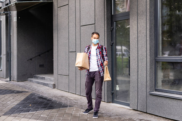 Un joven repartidor con máscara médica en la cara sosteniendo bolsas de papel en las manos. Hipster mensajero hombre con bolsas de compras. Servicio de entrega en línea seguro. Protección contra el Coronavirus COVID-19 - Foto, imagen