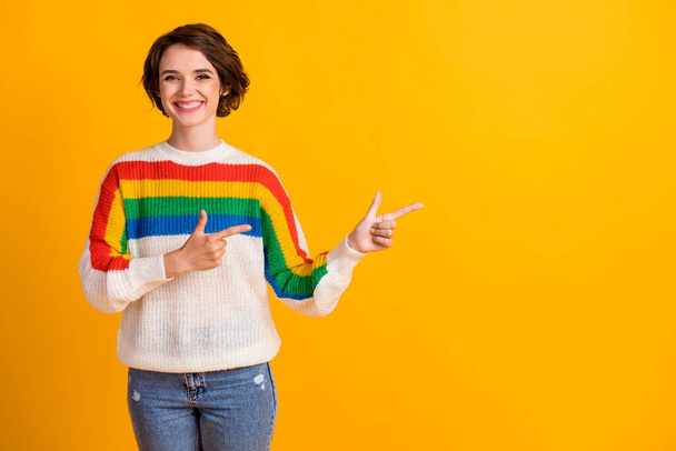 Photo of pretty smiling brunette bob hairstyle girl stand point empty space wear rainbow sweater blue jeans isolated on yellow background - Fotografie, Obrázek