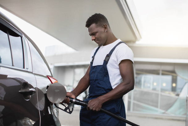 Petrol station, transportation and refueling concept. Handsome young black man, gas station worker, wearing dark blue overalls, refueling a car - Фото, изображение