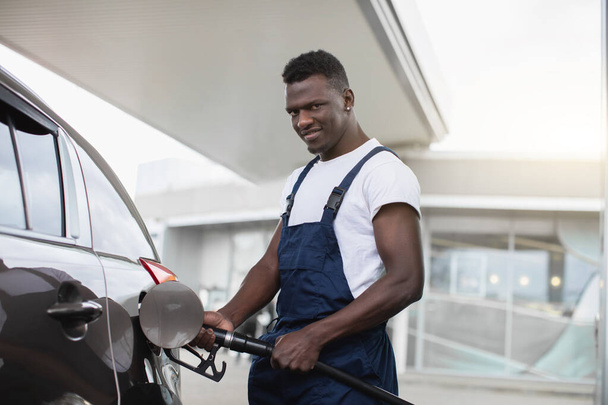 Petrol station, transportation and refueling concept. Handsome young black man, gas station worker, wearing dark blue overalls, refueling a car and looking at camera - Photo, image