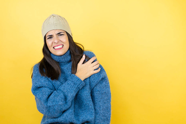Junge schöne Frau in blauem Pullover und Wollmütze mit Schmerzen auf der Schulter und schmerzhaftem Gesichtsausdruck - Foto, Bild