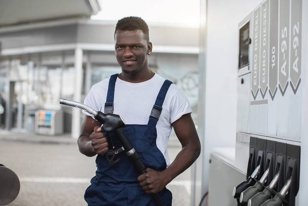 Young African man in workwear, worker of petrol station, is ready to refueling the car, holding filling gun nozzle, looking at the camera with smile - Foto, Bild