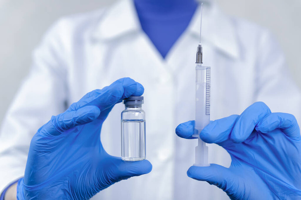 Kobieta w białym płaszczu, masce i niebieskich rękawiczkach trzyma strzykawkę w jednej ręce, a w drugiej butelkę ze szczepionką, zbliżenie leku. Koncepcja leku, leków i szczepień. - Zdjęcie, obraz