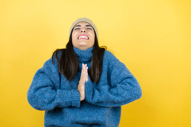 Fiatal gyönyörű nő kék alkalmi pulóvert visel és gyapjú kalap koldul és imádkozik kéz a kézben együtt remény kifejezés az arcon nagyon érzelmes és aggódik - Fotó, kép