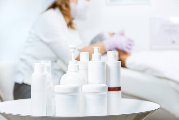 A set of cans and containers of skin care products in the foreground and a specialist beautician with a woman at the reception in the background. Mockup - Foto, Imagem